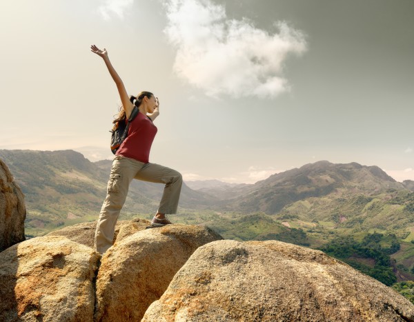 Hiker with backpack standing on top of a mountain with raised hands and enjoying landscape.